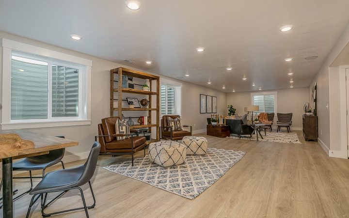 Fully finished basement recreation room with light wood laminate flooring, multiple seating areas including leather chairs and gray sofas, wooden bookshelf, patterned area rug, recessed ceiling lights throughout, and windows with white shutters