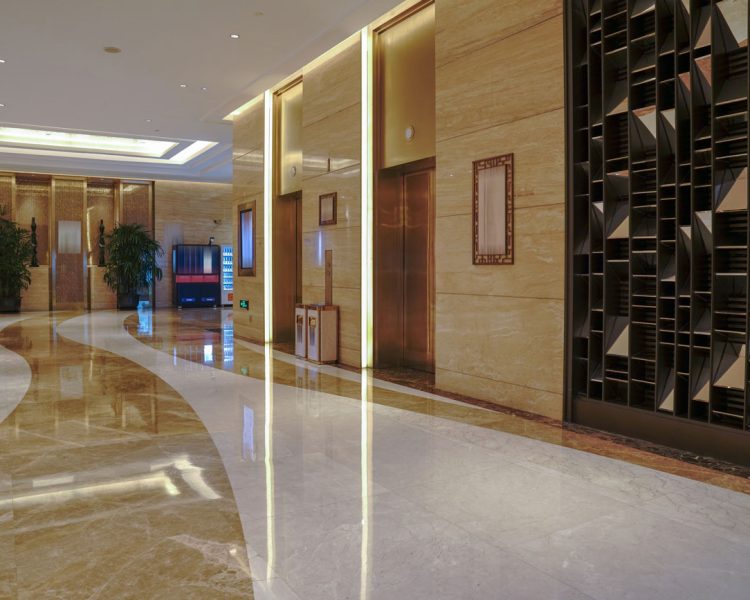 Elegant commercial lobby featuring polished marble floors with curved design patterns, golden-toned elevator doors with decorative lighting, beige stone walls, decorative geometric wall panels, and potted plants