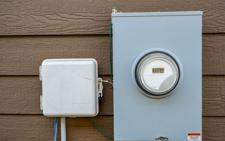 A modern digital electric meter and an adjacent utility box mounted on the brown siding of a house exterior.