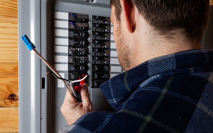 An electrician wearing a plaid shirt using wire strippers to prepare cables while working inside an open circuit breaker panel.