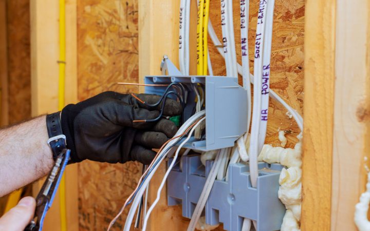 Close-up of electrician's gloved hands connecting wires in gray electrical junction boxes mounted on wooden wall studs, with labeled Romex cables for various circuits including LED, porch, and power