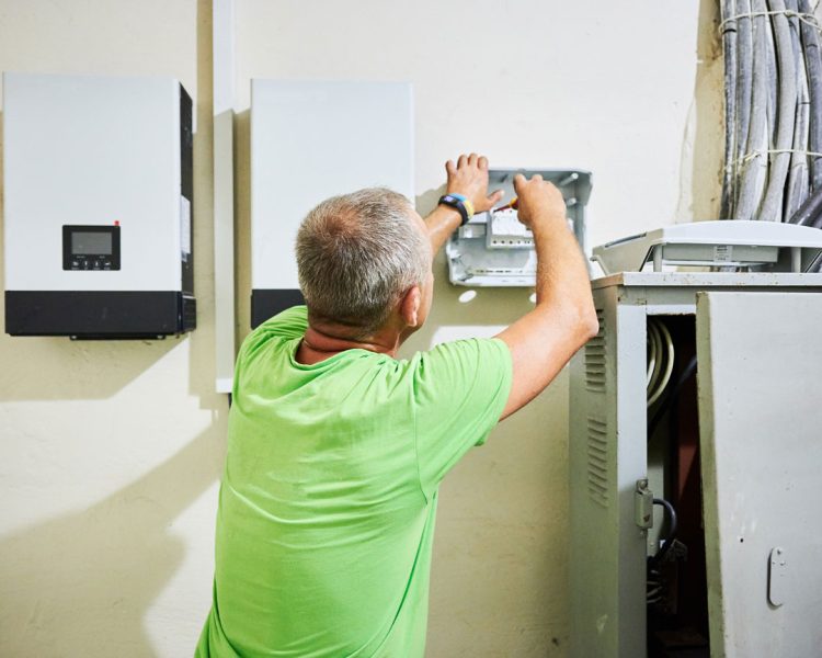 Electrician in bright green shirt installing or servicing wall-mounted solar inverters and electrical distribution equipment with exposed wiring and service panel