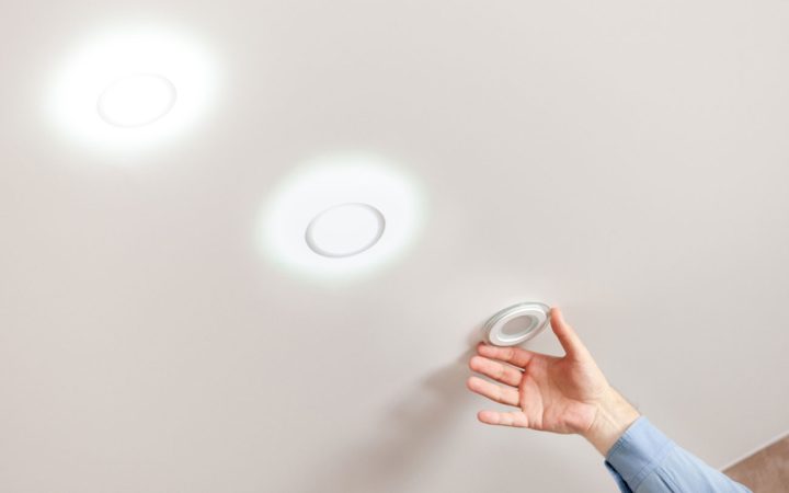 A hand holding a new, unlit LED wafer-style recessed light fixture up to a ceiling where two other identical fixtures are already installed and illuminated, demonstrating a modern lighting upgrade.