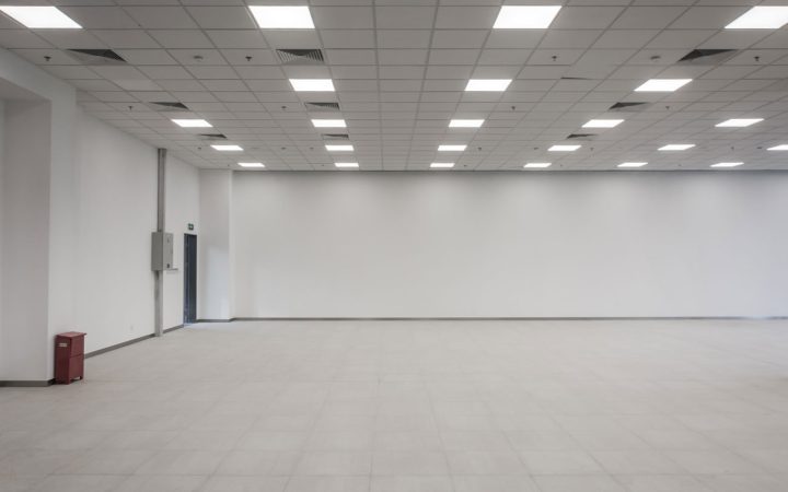 Spacious empty commercial interior featuring a suspended grid ceiling with numerous square LED panel lights, white walls, light tile flooring, and a fire extinguisher cabinet.