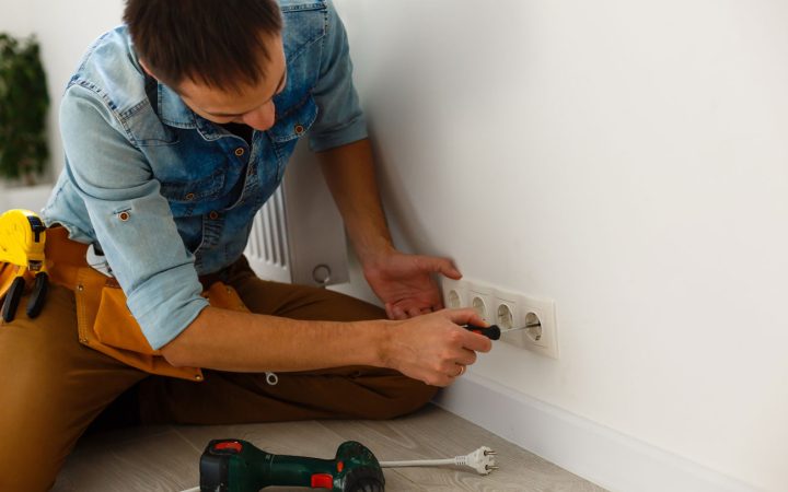 A handyman in a denim shirt kneels on the floor, using a screwdriver to install a series of white European-style electrical outlets on a clean white wall.