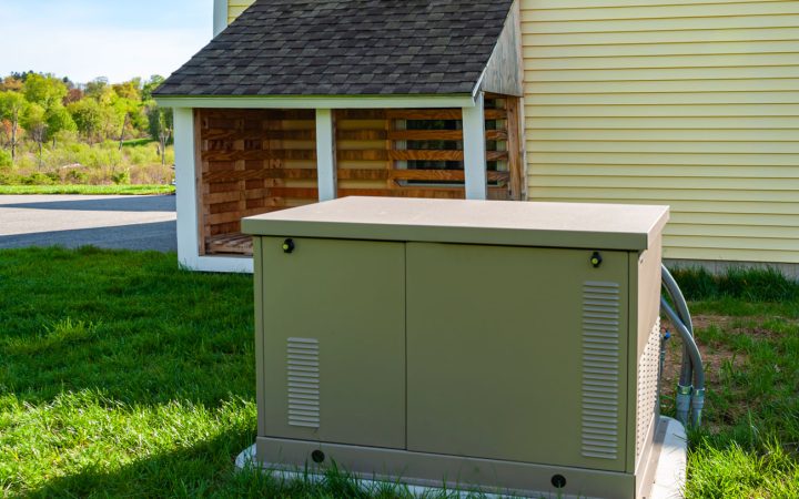 Tan residential standby generator installed on a concrete pad next to a yellow-sided house, with a wooden shelter structure nearby, set on a green lawn with trees in the background Filename: generator