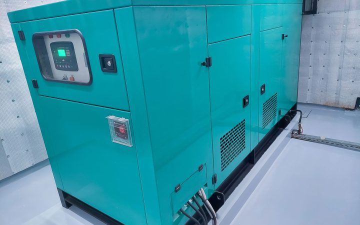 Large teal-colored commercial backup generator installed in a mechanical room, featuring a digital control panel display, ventilation grilles, and professional indoor installation with gray flooring