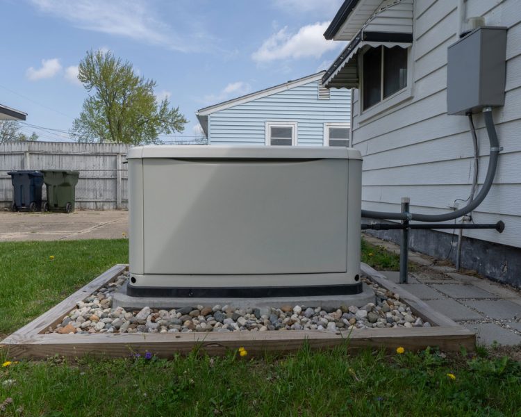 Beige whole-house standby generator installed on a concrete pad surrounded by decorative river rocks in a wooden frame, positioned next to a white vinyl-sided home with electrical conduit connection