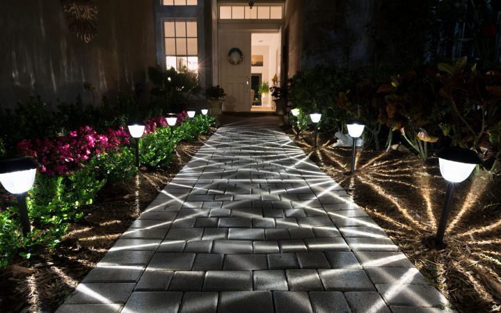 A paved walkway leading to a front door illuminated by path lights that cast dramatic starburst shadows across the bricks and surrounding mulch beds.